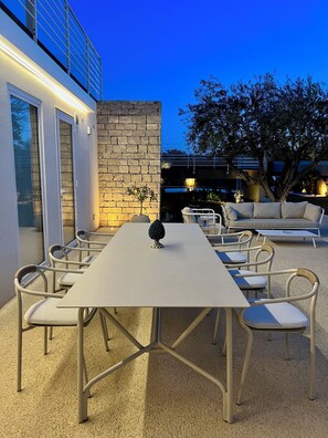 An outdoor patio with a long table and chairs, a small tree, and a sofa.