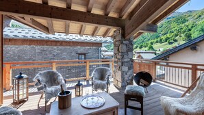 Eine Holzterrasse mit einer Steinmauer, zwei weiße Stühle, ein Holztisch mit einer Flasche und Gläsern sowie eine Steinsäule.