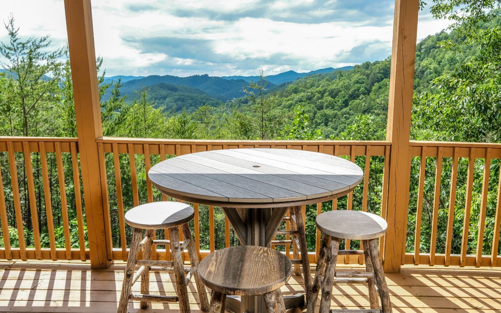 A wooden deck with a round table and two stools, overlooking a mountainous landscape.