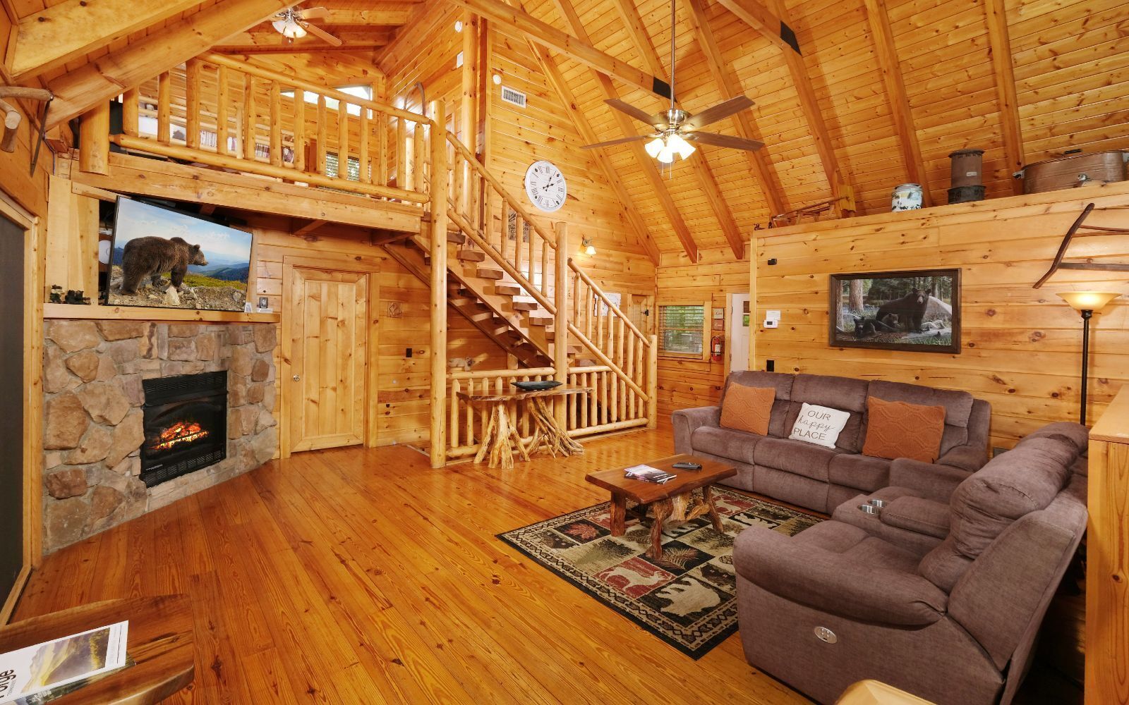 A wooden cabin interior with a stone fireplace, a wooden staircase, a living room with a sofa and a rug, and a ceiling fan.