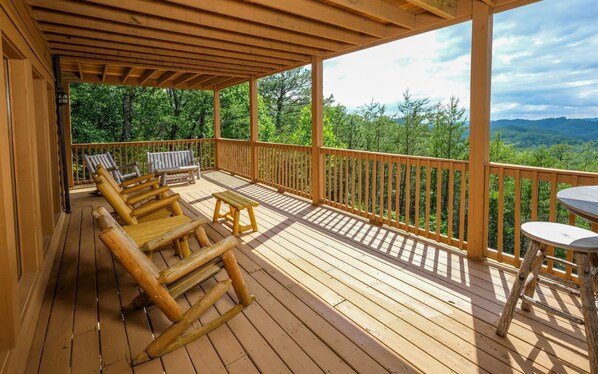 A wooden deck with outdoor seating, including chairs and a table, overlooking a forested landscape. - Cali's Place - Gorgeous Mountain Views, Pet Friendly, Hot Tub + Pool Table (Sevierville)