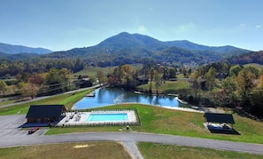 A swimming pool with a deck, surrounded by a parking area and two buildings, set against a backdrop of mountains and autumn foliage.