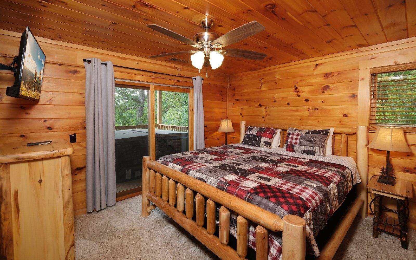 Une chambre dans une cabane en bois, avec un lit, un ventilateur de plafond, une fenêtre avec des rideaux et une petite table avec une lampe.