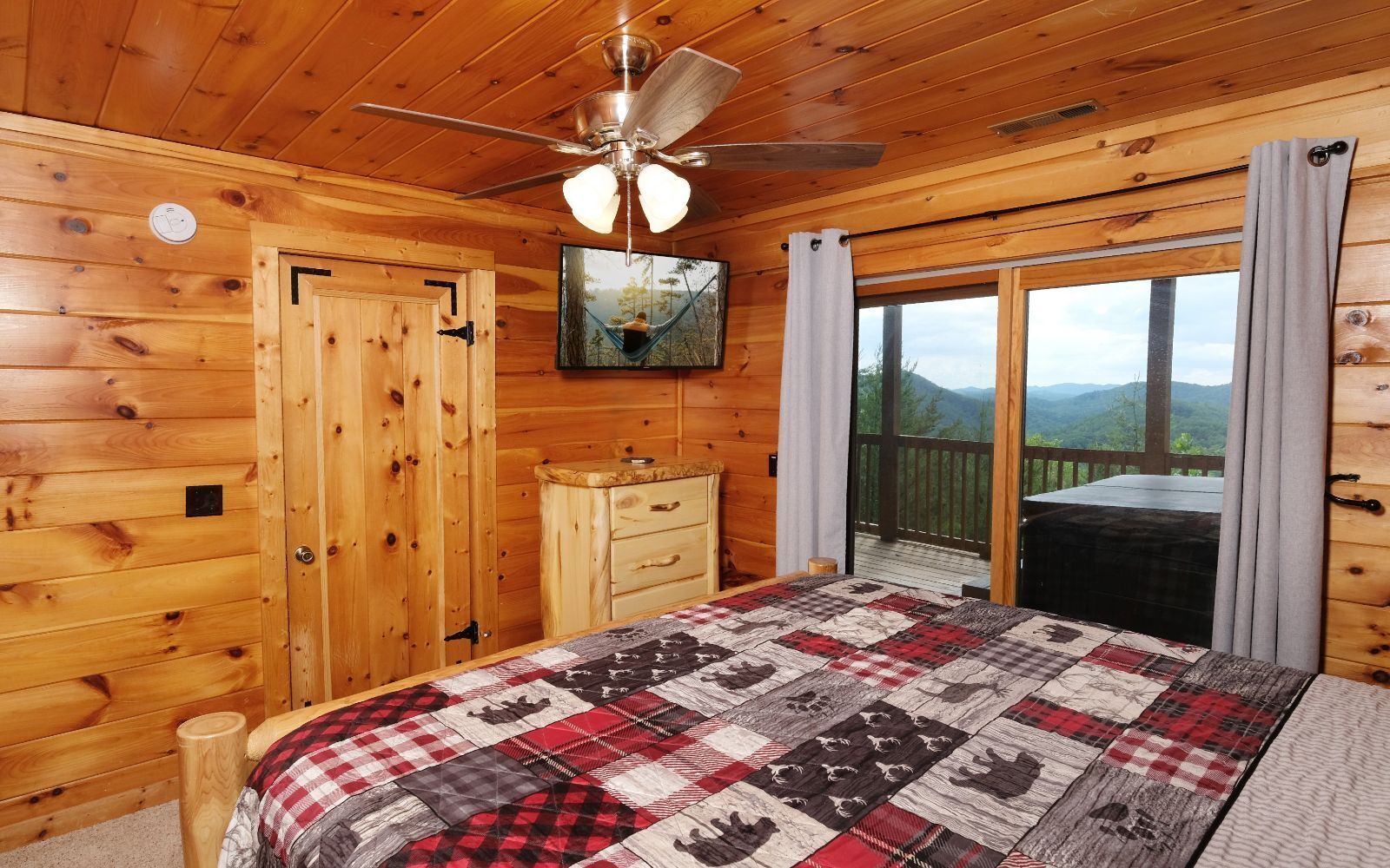Une chambre à coucher aménagée dans une cabane en bois, avec un lit, une commode, un ventilateur de plafond et une vue sur les montagnes, visible à travers une porte-fenêtre coulissante.