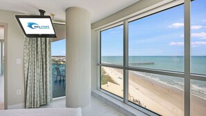 Une chambre dotée d’une grande fenêtre donnant sur une plage, d’un téléviseur à écran plat fixé au mur et d’un rideau à motifs.