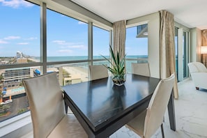 A hotel room with a large window offering a view of the beach and cityscape, a dining table with chairs, and a potted plant.