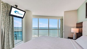 Une chambre d’hôtel avec un grand lit, un téléviseur fixé au mur et une vue sur l’océan grâce à de grandes fenêtres.
