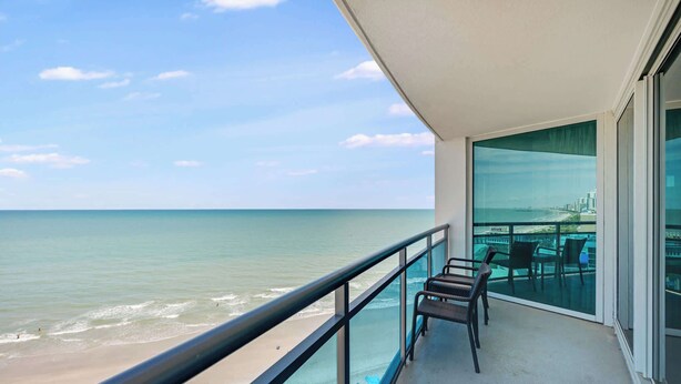 Un balcon avec vue sur la plage et l’océan, une porte vitrée et un espace salon avec des chaises.