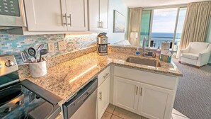 A modern kitchen with granite countertops, white cabinets, and a coffee maker. There is a view of the ocean through large windows. - New Top Floor Direct Oceanfront with Amazing View! (Myrtle Beach)