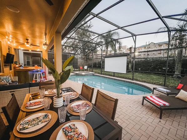 An outdoor pool area with a dining setup, including a table with plates and glasses, and a bar area with a TV.