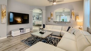 A modern living room with a large flat-screen TV, a white sofa, a coffee table, and a ceiling fan. - Luxury 6BR | Game Room, Outdoor Theater & Pool/Spa (Kissimmee)