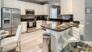 A modern kitchen with white cabinets, stainless steel appliances, a granite countertop, and a dining area with white chairs. - Luxury 6BR | Game Room, Outdoor Theater & Pool/Spa (Kissimmee)