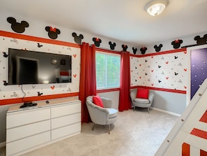 A room with a TV, two chairs, a white dresser, and Mickey Mouse wall decals.