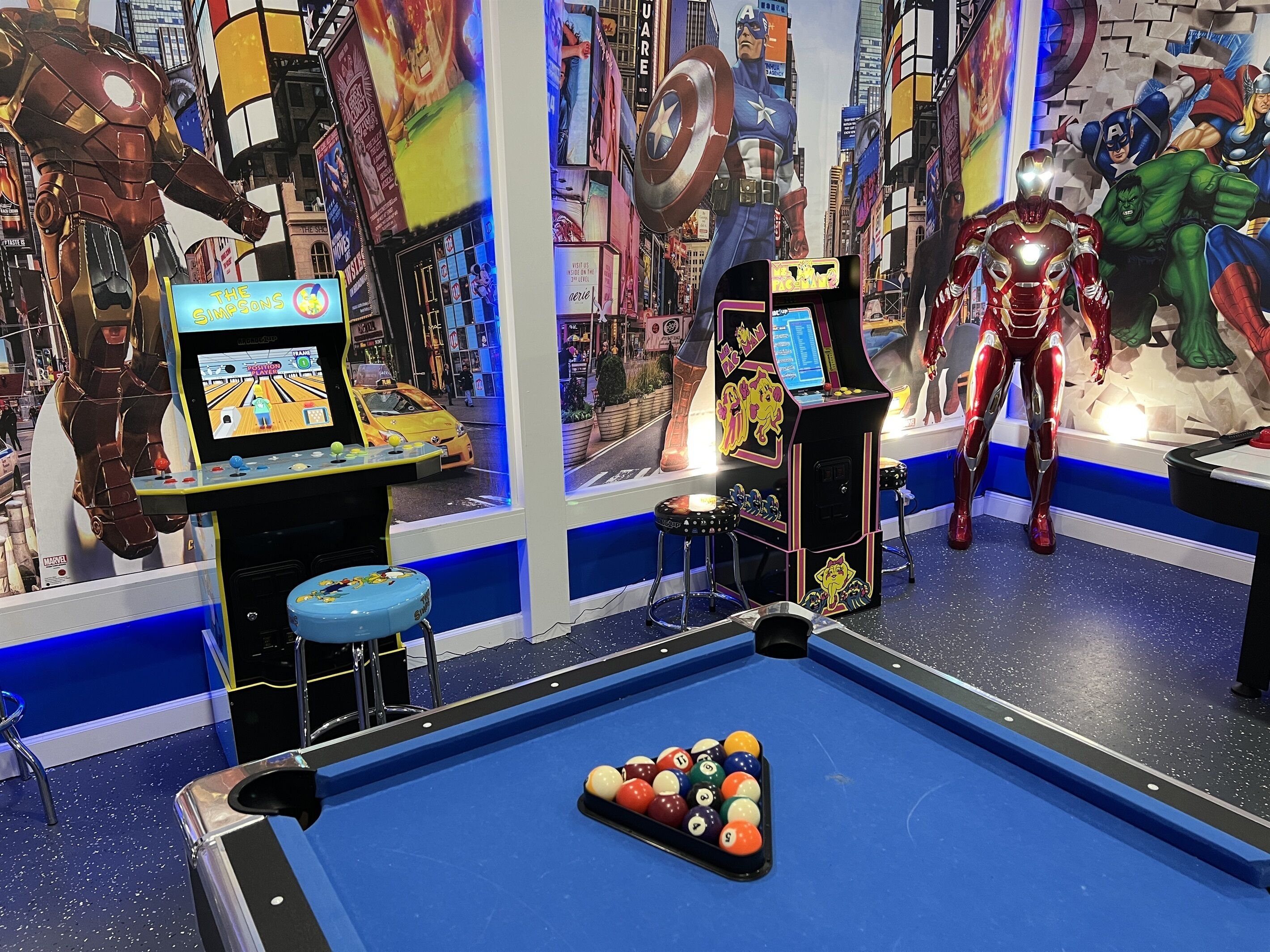 A pool table with a pool cue, a bowling alley, and arcade machines with superhero themes.