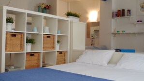 Een slaapkamer met een bed, witte muren, planken met spullen en rieten manden.