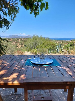 Un tavolo di legno con un bicchiere sopra, posizionato su un patio con vista su un paesaggio pittoresco, montagne e uno specchio d'acqua in lontananza.