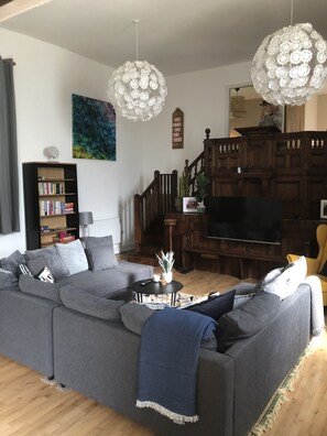 Ein modernes Wohnzimmer mit einem grauen Ecksofa, einer Holztreppe, einem Bücherregal und einem großen Flachbildfernseher.