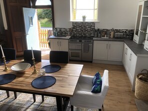 Eine moderne Küche mit weißen Schränken, einem eingebauten Ofen und einem Herd. Es gibt eine Essecke mit einem Holztisch, Stühlen und einer blauen Matte. Ein Fenster lässt natürliches Licht herein, und eine Tür führt in einen Außenbereich.
