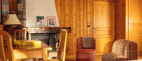 A cozy room with wooden walls, a fireplace, a dining table with chairs, and a plaid armchair.