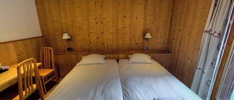 A room with two beds, a wooden desk, two chairs, and a window with curtains.