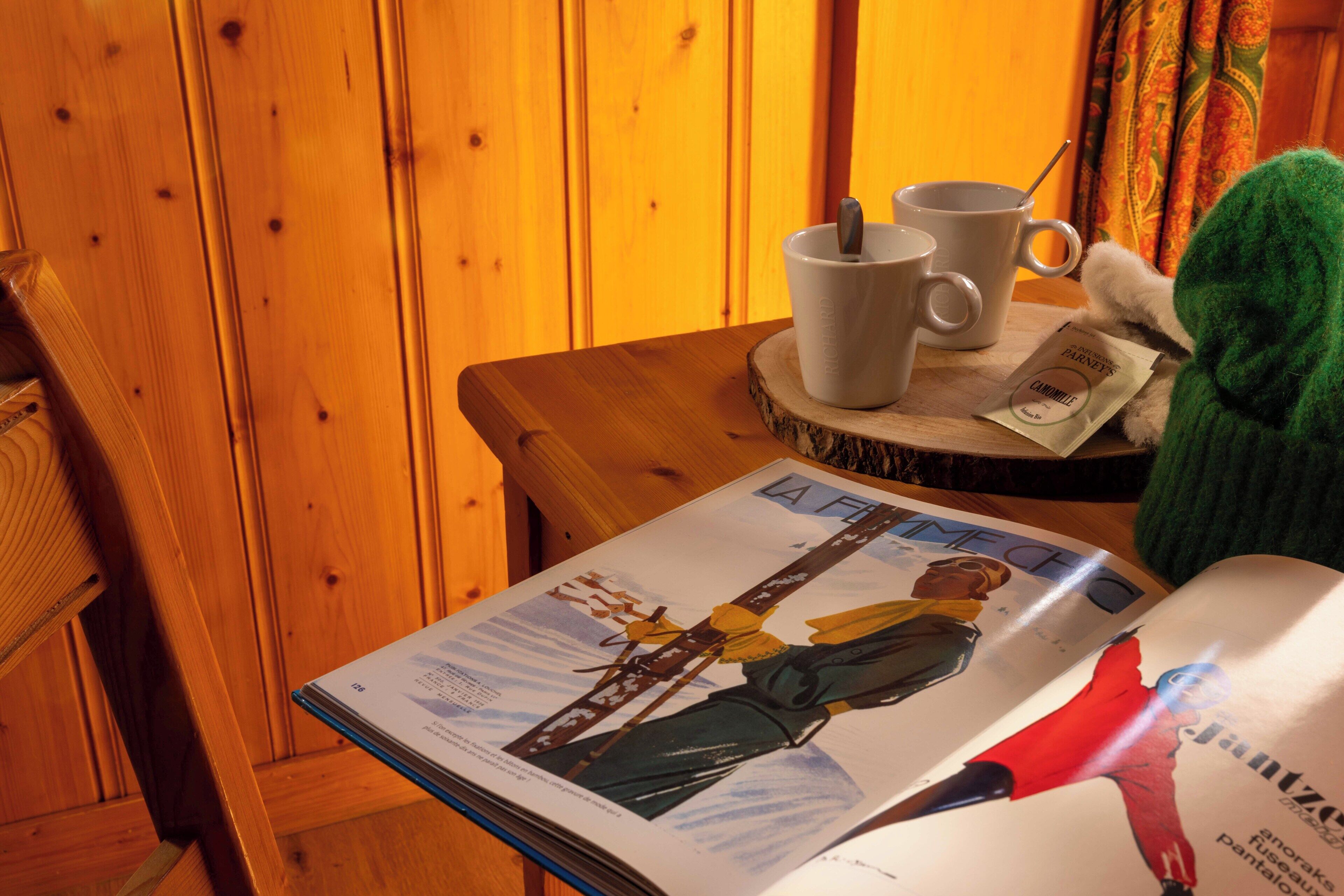 A magazine on a wooden table with two white mugs and a spoon, a green plush toy, and a wooden chair.