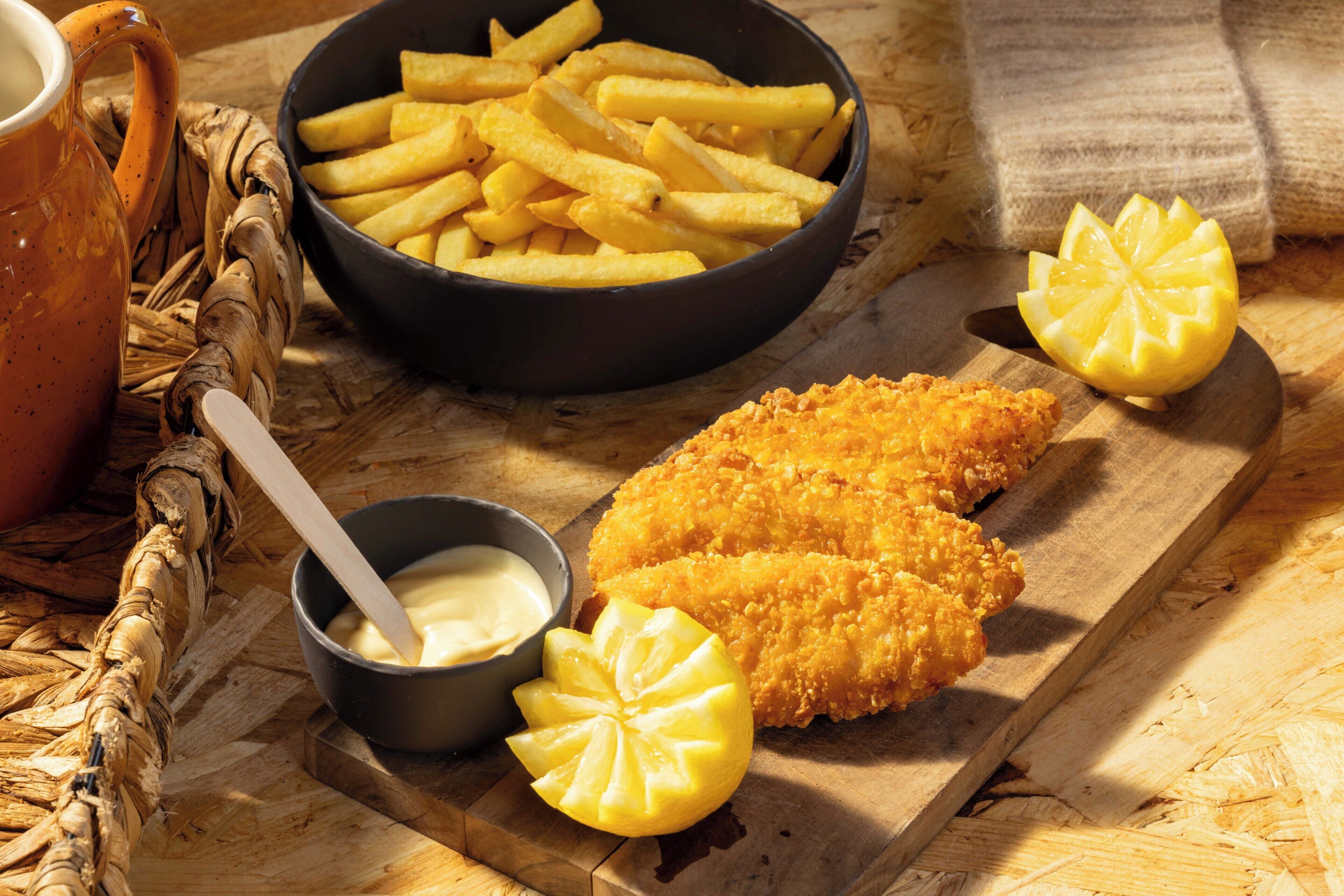 Una tabla de madera con pescado rebozado, un bol de papas fritas, una rodaja de limón y un recipiente pequeño con salsa.