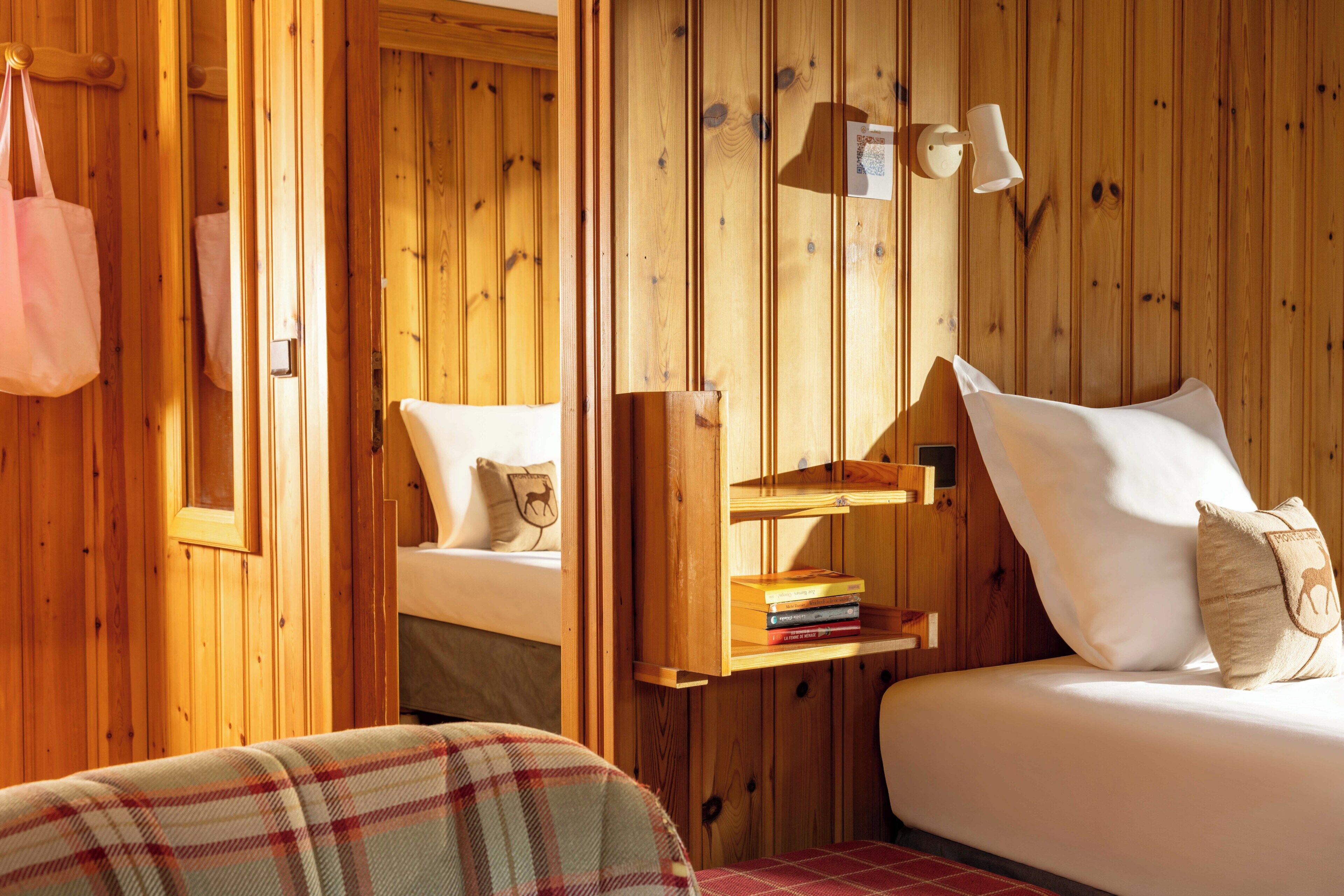 A wooden room with a bed, a shelf with books, a hanging bag, and a wall-mounted lamp.