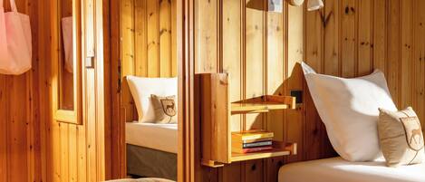 A wooden room with a bed, a shelf with books, a hanging bag, and a wall-mounted lamp.