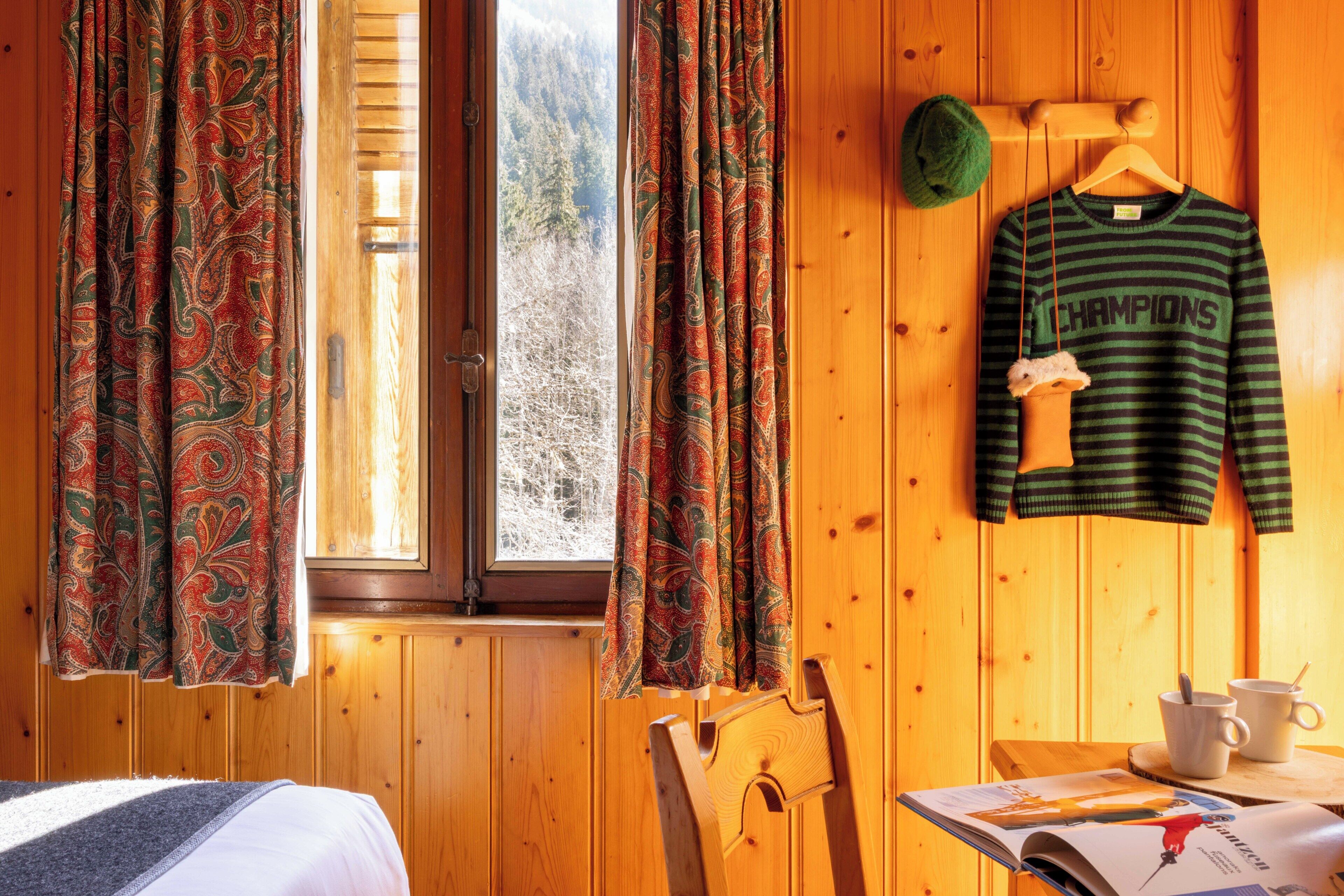 A wooden room with a bed, a chair, a table with a magazine, a sweater, and curtains.