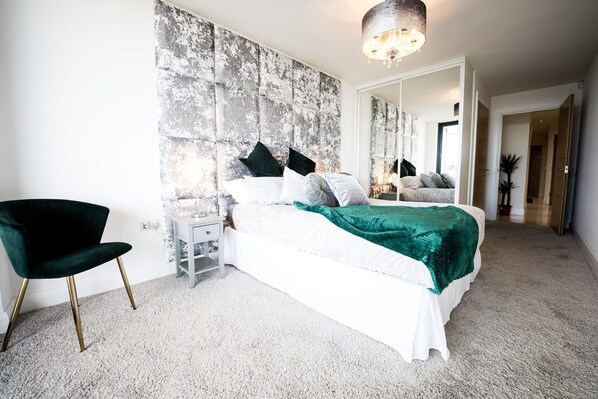 A bedroom with a bed, a nightstand, a chair, a large mirror, and a chandelier. - No2 Sandy Bay , Luxury Beachfront Apartment (Portrush)