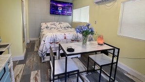 A bedroom with a bed, a TV, an air conditioner, a table with chairs, and a window with blinds.