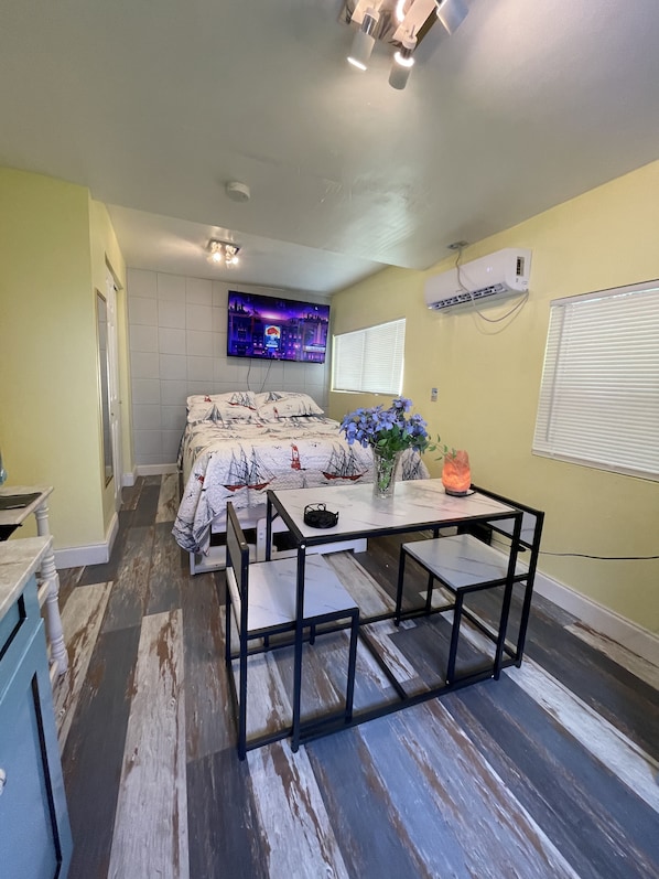 A bedroom with a bed, a TV, an air conditioner, a table with chairs, and a window with blinds.