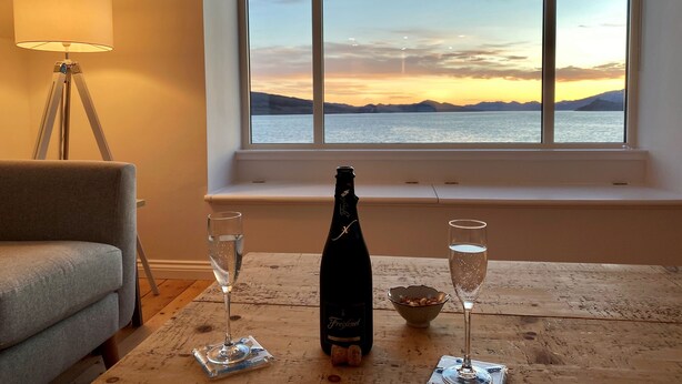 A room with a view of a sunset over water, a bottle of champagne, and two glasses on a wooden table. - Greenlea -Scenic loch-side location, coastal retreat boasts spectacular sea views, sleeps 4 (Strachur)