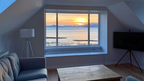 A room with a large window overlooking a sunset over water, a tripod lamp, a sofa, and a wooden table. - Greenlea -Scenic loch-side location, coastal retreat boasts spectacular sea views, sleeps 4 (Strachur)