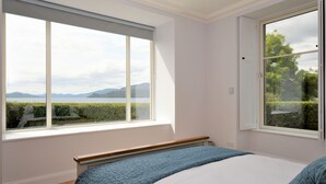 A bedroom with a large window, a bed with blue bedding, and a view of a landscape with mountains and trees. - Greenlea -Scenic loch-side location, coastal retreat boasts spectacular sea views, sleeps 4 (Strachur)