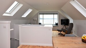 A modern loft bedroom with skylights, a large window with a view, a flat-screen TV, and a wooden dining table. - Greenlea -Scenic loch-side location, coastal retreat boasts spectacular sea views, sleeps 4 (Strachur)