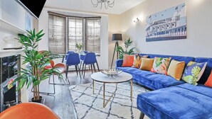 A living room with a blue sofa, a round coffee table, and a chandelier. There are potted plants and a painting on the wall. - Central Lanes Pad | By My Getaways | Free parking for one car (Brighton)