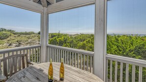 A wooden deck with a table and two bottles of wine, overlooking a beach with greenery and a distant view of the ocean.