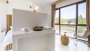 A modern hotel room with a large window, a bed, a small table with a bowl of fruit, and a chair.