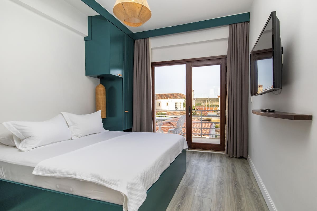 Habitación de hotel con cama, televisión, balcón con vistas a edificios y suelo de madera.