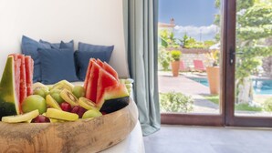 A wooden bowl filled with assorted fruits on a table, with a view of a pool and greenery outside a room with blue pillows.