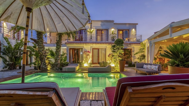 A poolside area with lounge chairs, a large umbrella, and a building with balconies and potted plants.