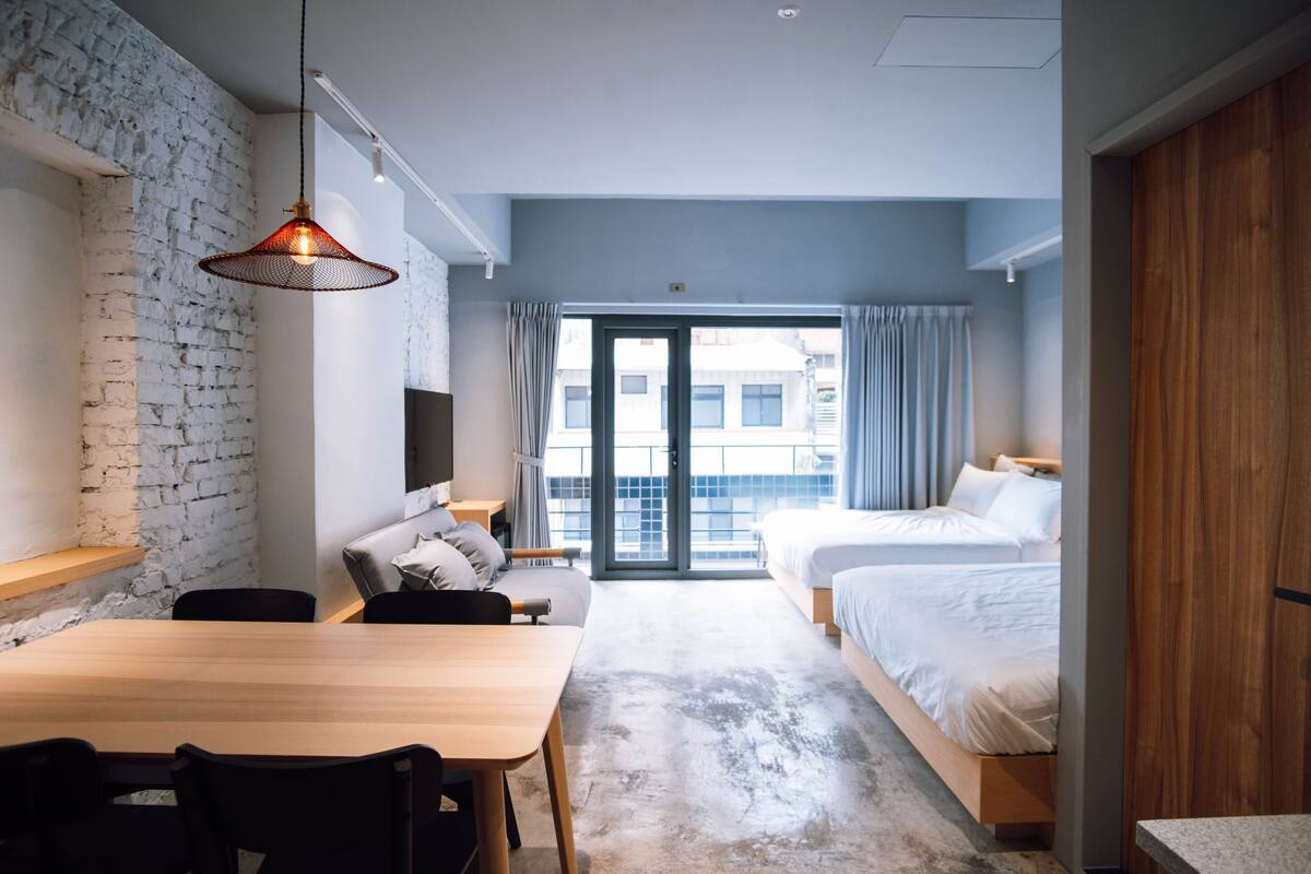 Una habitación de hotel moderna con una mesa de comedor de madera, dos camas, un amplio ventanal con cortinas y una pared de ladrillo.