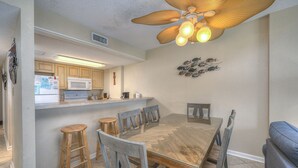 A dining area with a wooden table and chairs, a kitchen in the background, and a ceiling fan with lights. - Bluewater Resort 901 - 2 Bedroom/Bath Ocean View Condo, Indoor/Outdoor Pool, Hot tub (Myrtle Beach)
