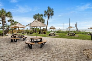 Un espace extérieur aménagé avec une surface pavée, des tables de pique-nique et des parasols, des palmiers, et une vue sur l’eau.