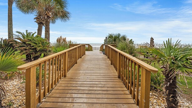 Un paseo marítimo de madera que conduce a una playa con palmeras y cielo despejado.