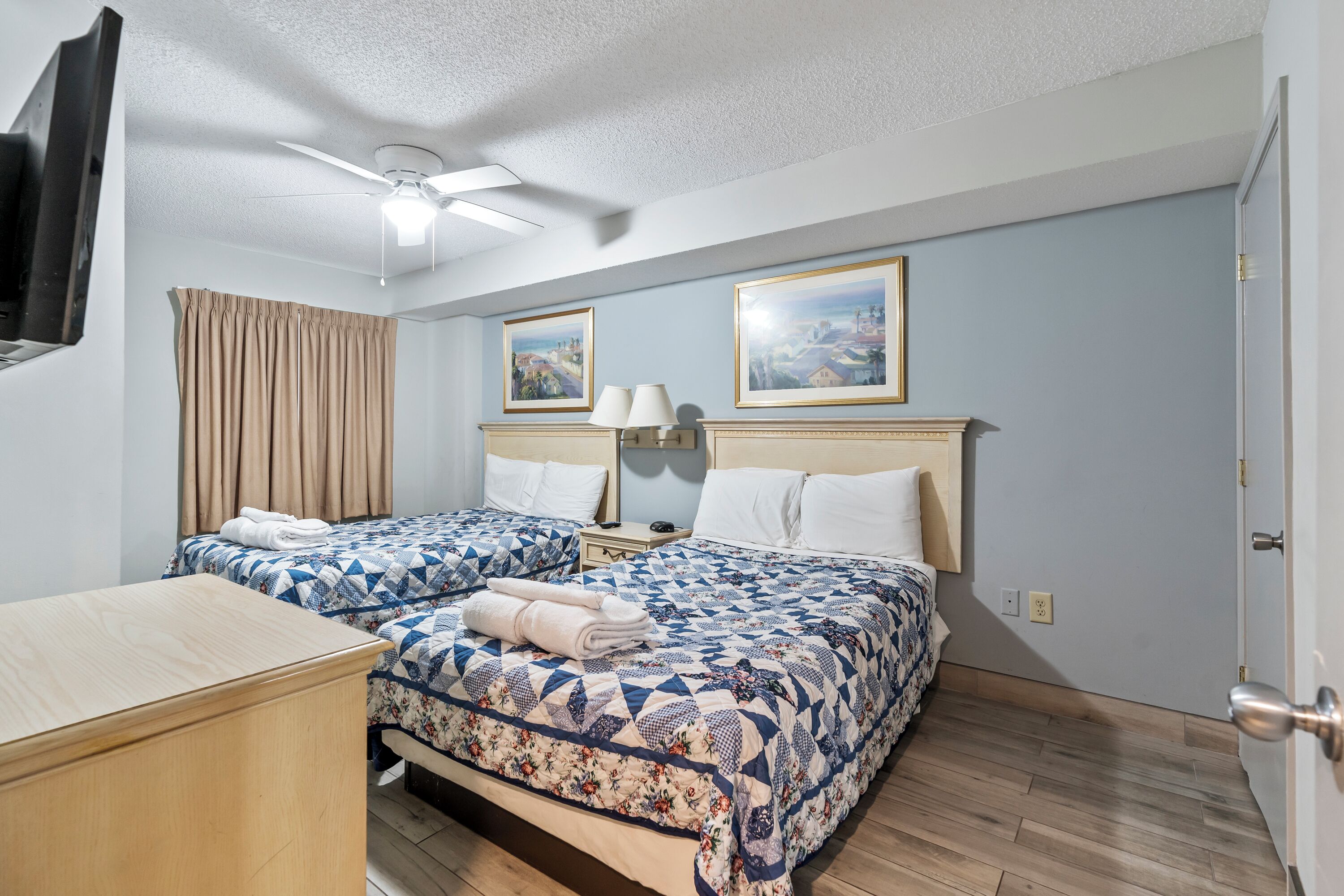 Een hotelkamer met twee bedden, een houten dressoir, een plafondventilator en twee ingekaderde foto's aan de muur.