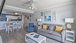 A modern living room with a grey sofa, a blue armchair, and a white coffee table. There is a dining area with white furniture and a kitchen in the background.