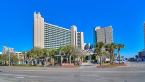 Um hotel imponente com vários andares, entrada destacada e área de estacionamento com uma van branca.