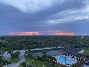 Une propriété résidentielle dotée d’une piscine, d’un court de tennis et offrant une vue sur le coucher de soleil se reflétant sur un paysage boisé.